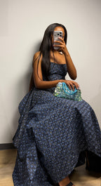 A woman takes a mirror selfie wearing a dark navy blue patterned strapless two piece,with a hertunba inspired voluminous skirt, holding a patterned clutch bag.