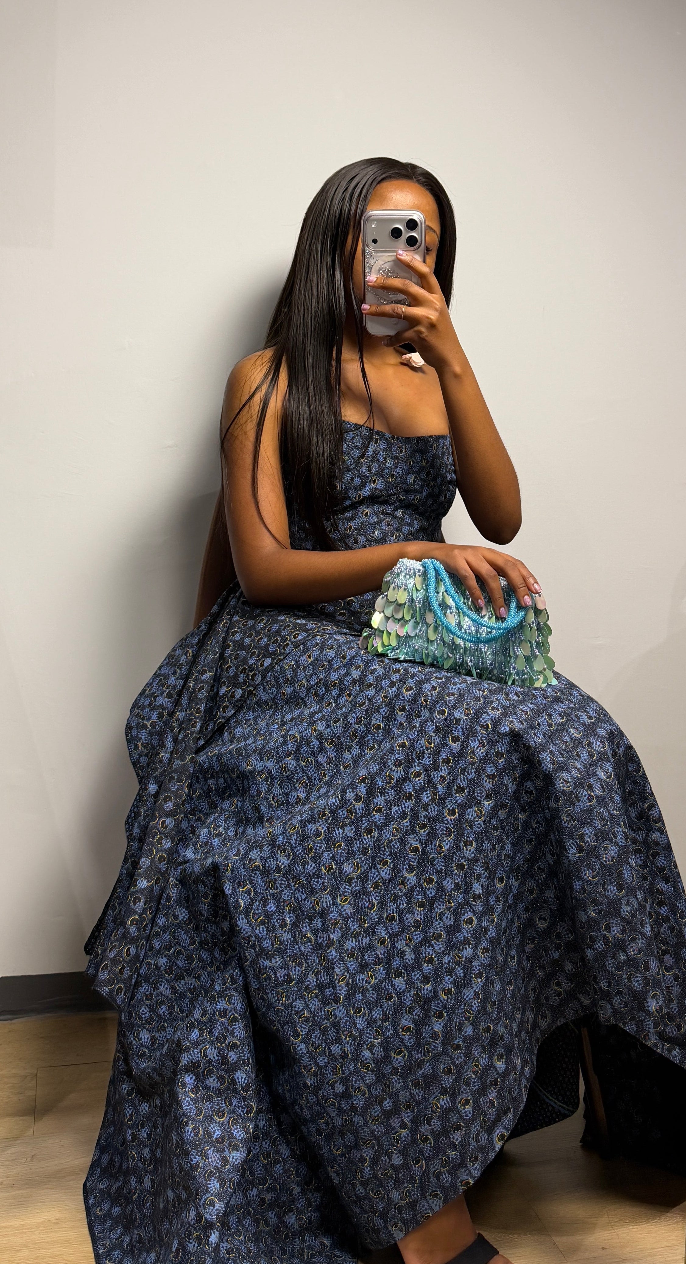 A woman takes a mirror selfie wearing a dark navy blue patterned strapless two piece,with a hertunba inspired voluminous skirt, holding a patterned clutch bag.